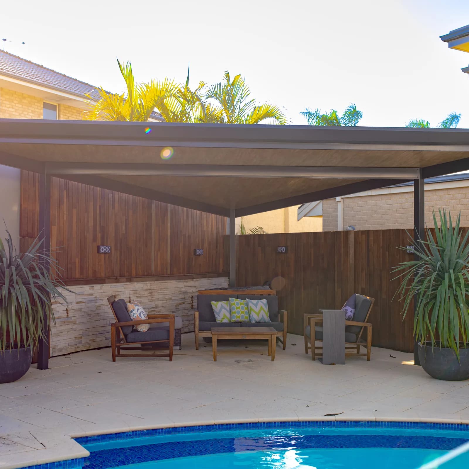 A wide flat roof patio beside a pool, covering an outdoor lounge setting against a timber fence