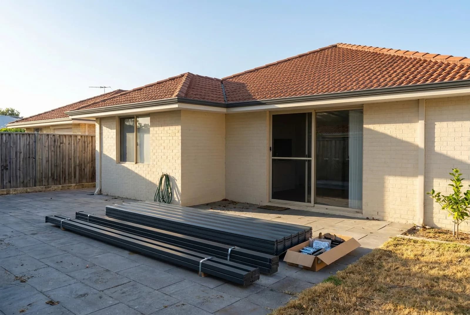 A flat-packed steel kit laid out on the paving behind a Perth home, beams and posts strapped in bundles beside a box of brackets and bolts