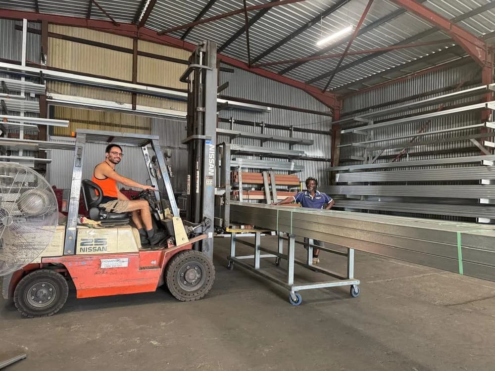 A steel patio frame on a trolley being moved through The Patio Factory's Welshpool workshop, with racked steel sections behind it